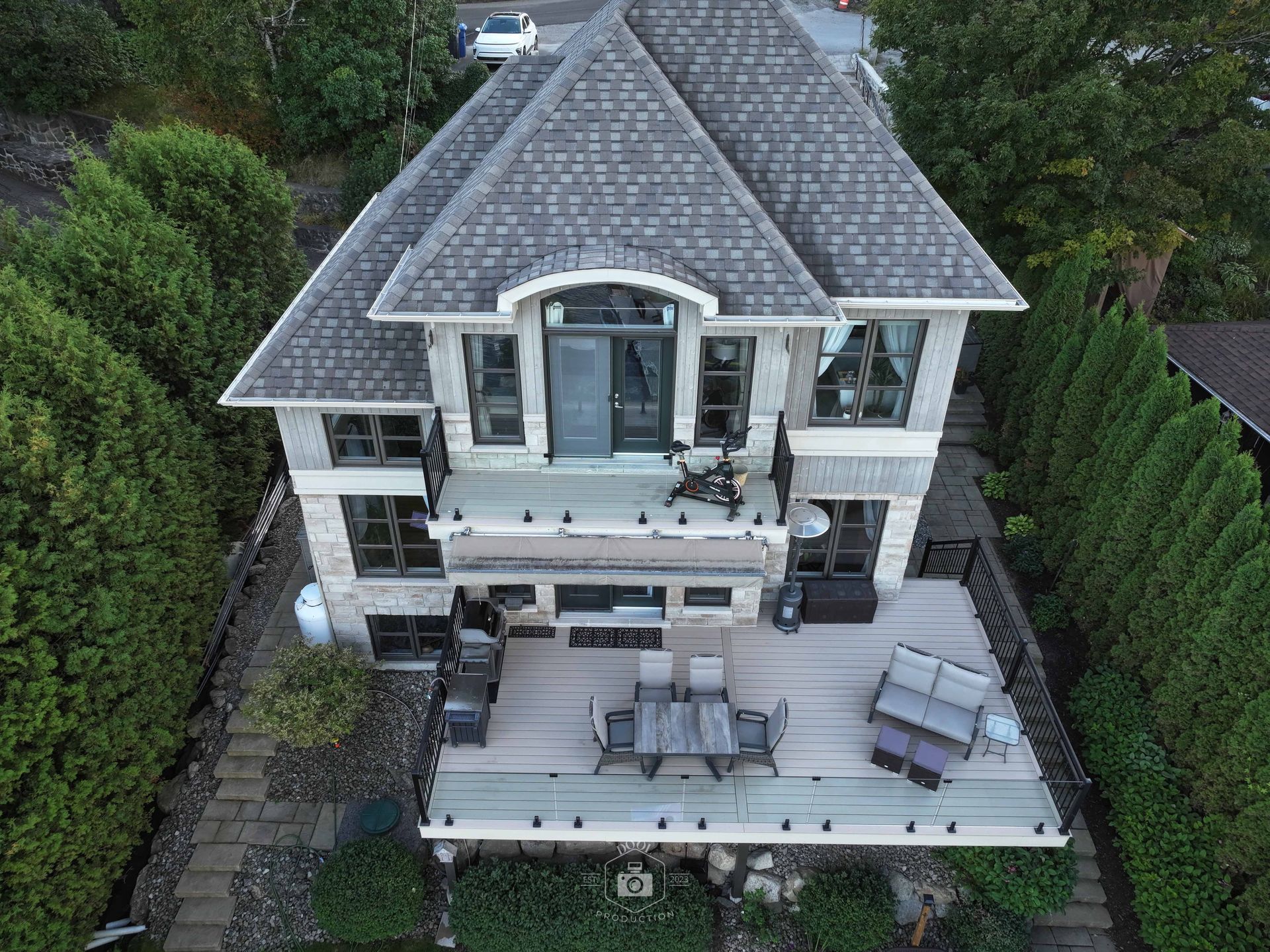 Maison en pierre de deux étages avec terrasse, entourée d'arbres. Toit gris, portes vitrées et mobilier de jardin.