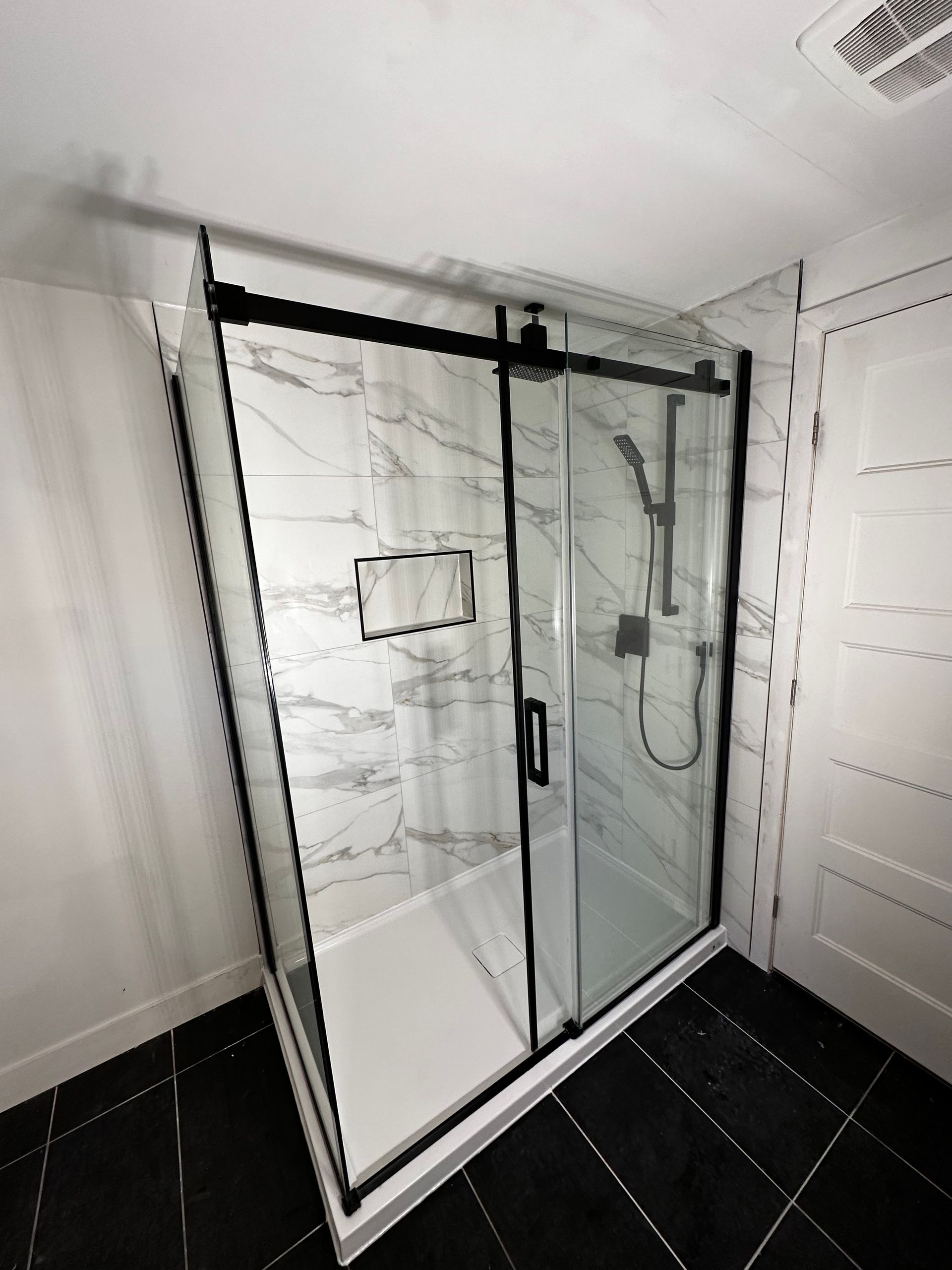 Douche moderne avec cadre noir, porte coulissante en verre, carrelage en marbre blanc et sol noir.