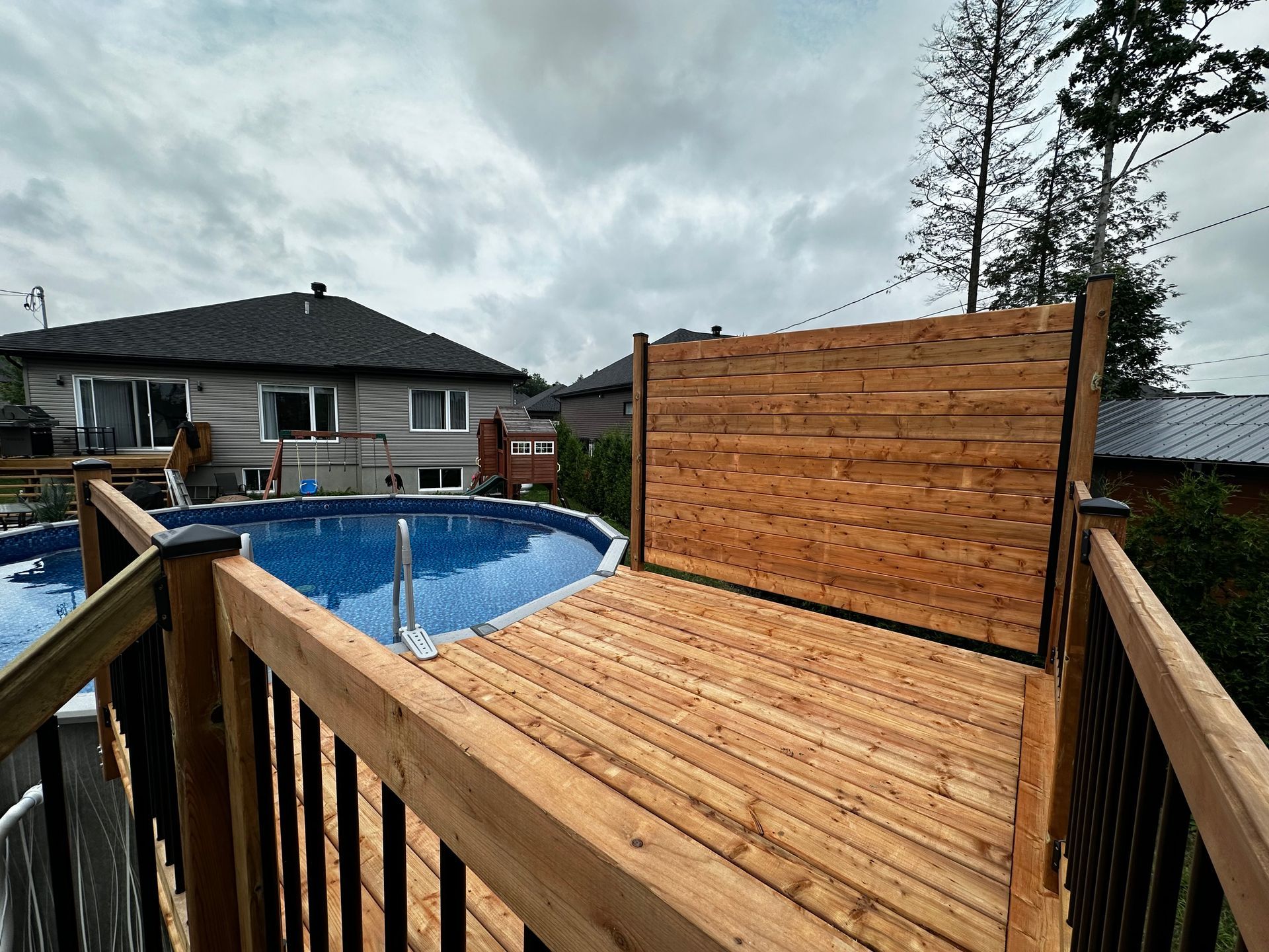 Terrasse en bois et paravent en cèdre pour plus d'intimité à côté d'une piscine hors sol sous un ciel nuageux.