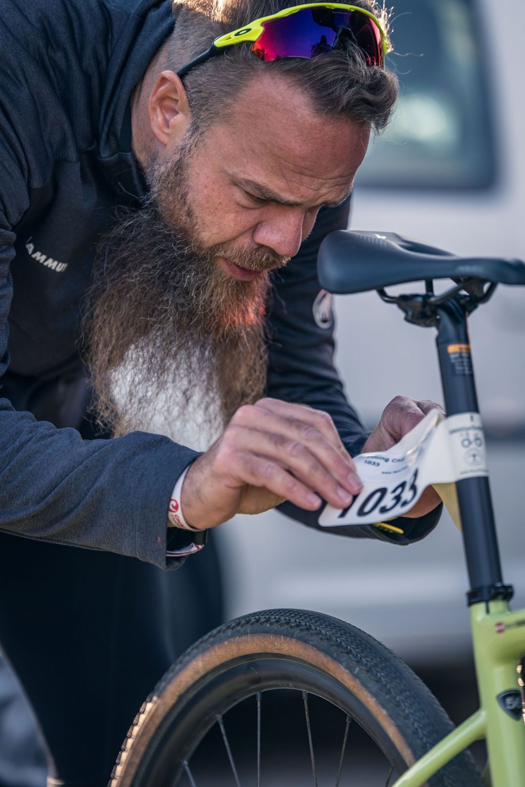 Ein Radfahrer befestigt im Freien eine Startnummer an seinem Fahrrad; Vorderrad und Lenker sind sichtbar.