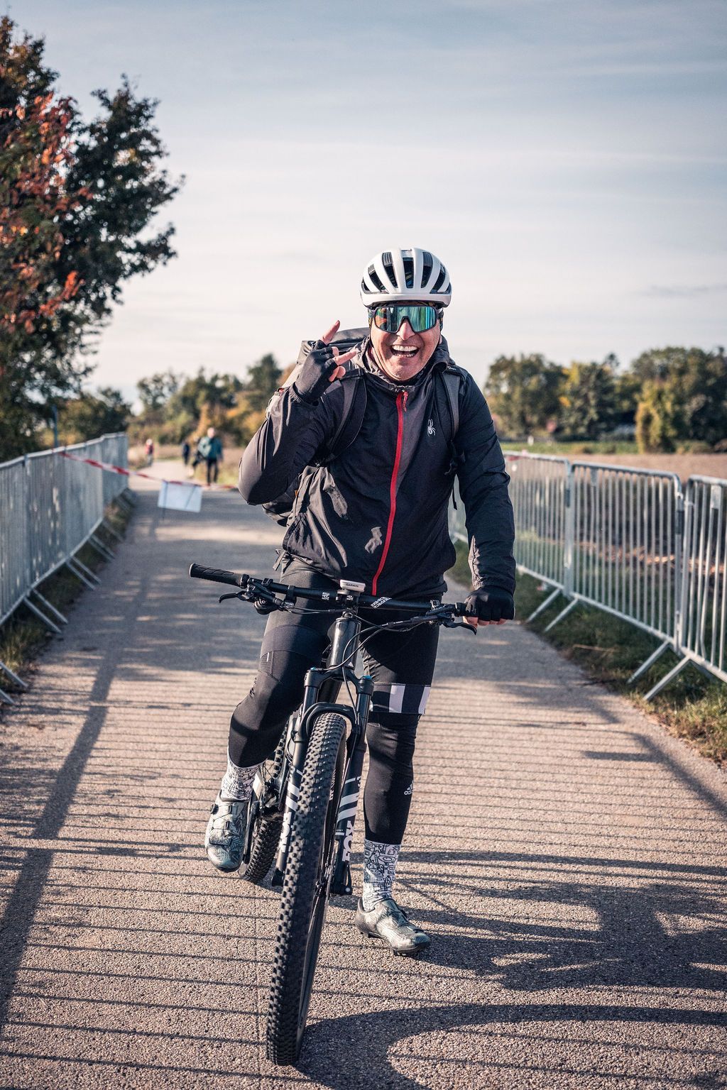 Ein Radfahrer mit Helm fährt auf einer Brücke, hebt zwei Finger und lächelt.