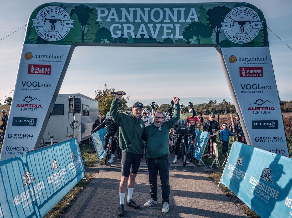 Zwei Radfahrer an der Ziellinie des Pannonia Gravel-Rennens, die mit erhobenen Armen unter einem Sponsorenbogen posieren.