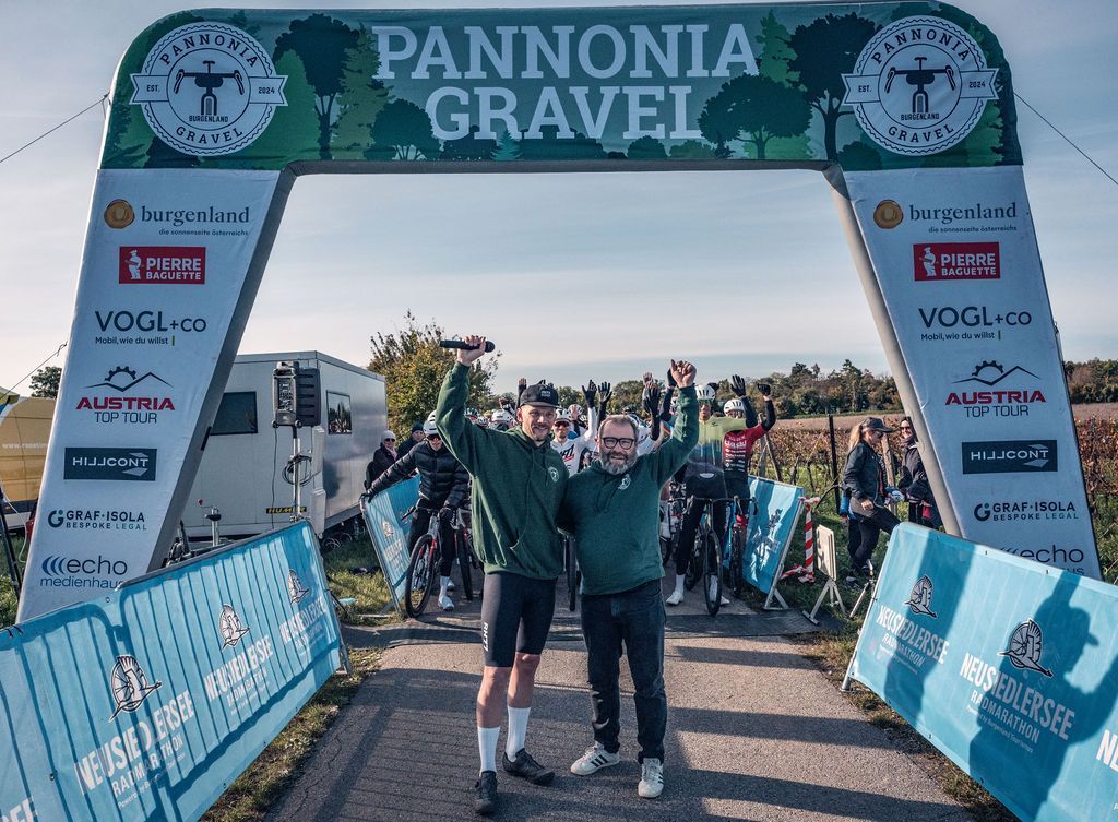 Zwei Radfahrer feiern unter dem Zielbogen des Pannonia Gravel-Rennens auf einer mit Absperrungen gesäumten Straße.