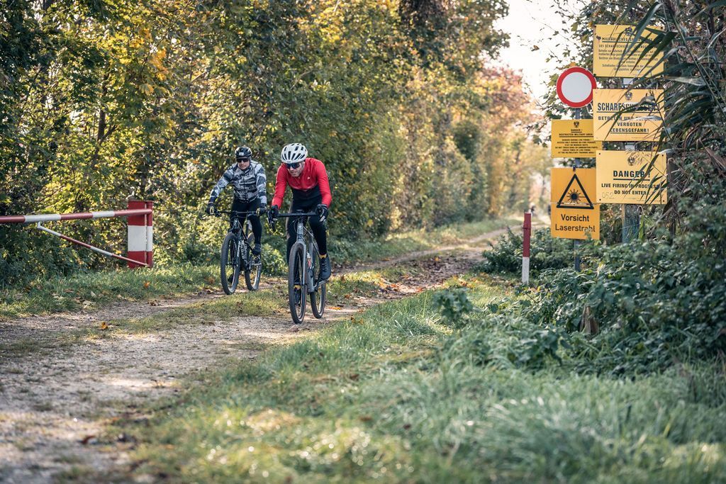 Zwei Radfahrer fahren auf einem unbefestigten Weg neben Warnschildern in einem Waldgebiet.