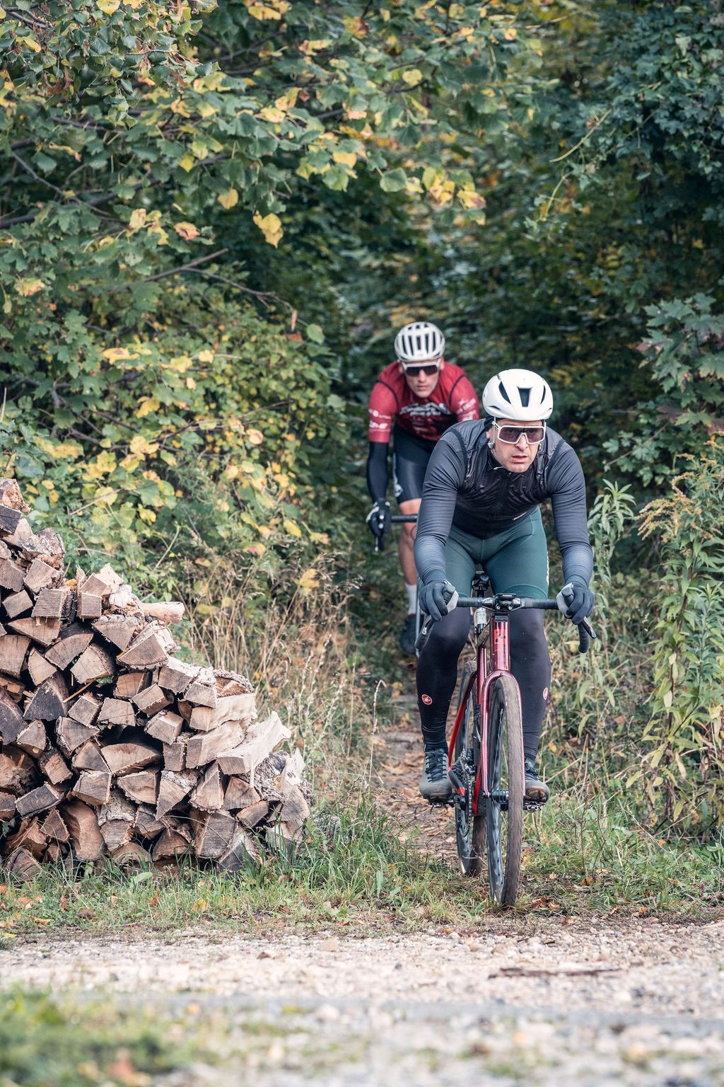 Zwei Radfahrer fahren hintereinander auf einem Feldweg neben gestapeltem Brennholz und dichtem Grün.