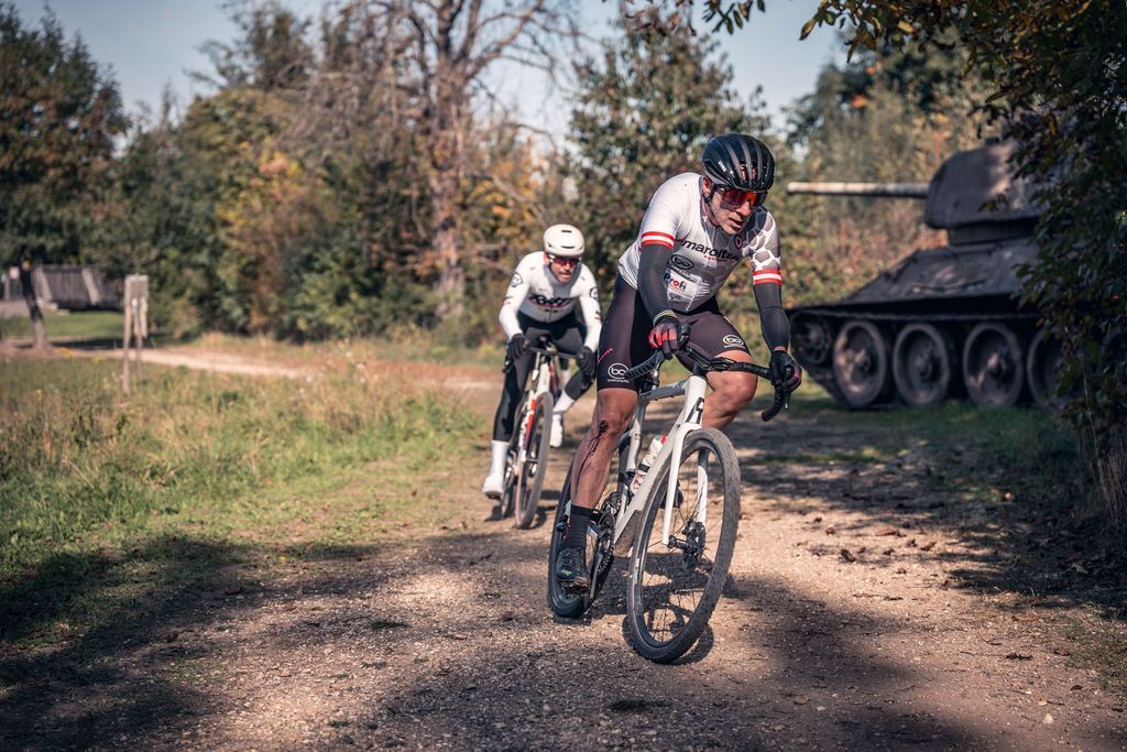 Drei Radfahrer liefern sich ein Rennen auf einem unbefestigten Weg neben einem geparkten Panzer in einem bewaldeten Feld.