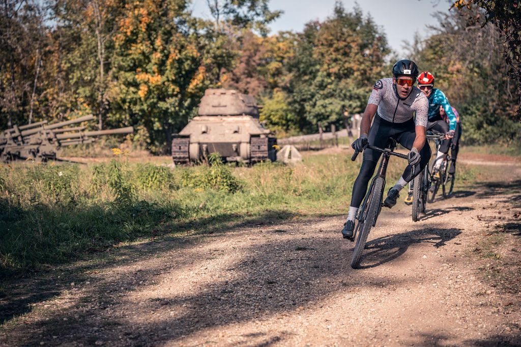 Radfahrer rasen auf einem Feldweg neben einem bewaldeten Feld; im Hintergrund ist ein kleiner Betonbunker zu sehen.