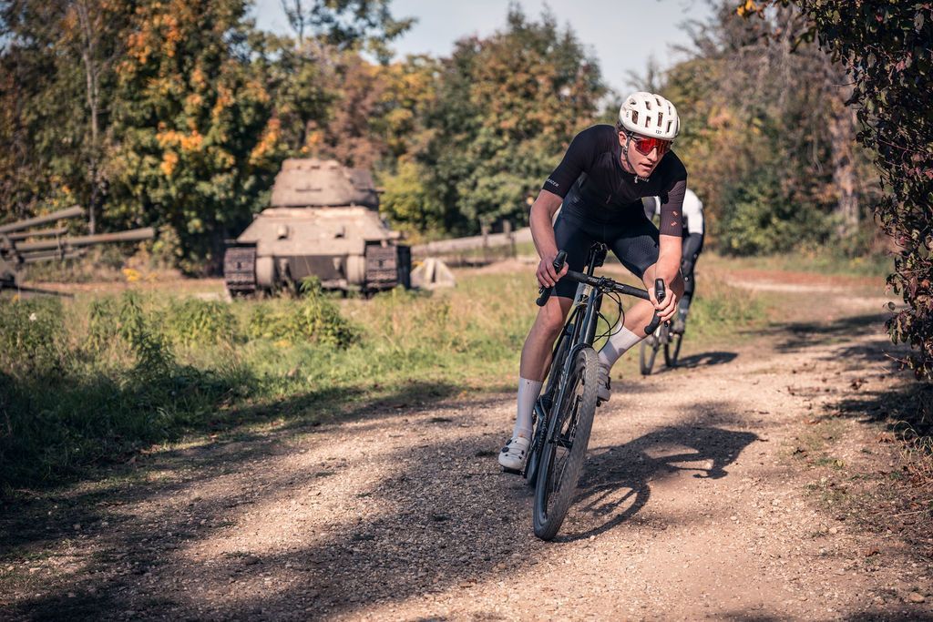 Ein Radfahrer in Schwarz fährt auf einem Schotterweg neben einem rustikalen Steingebäude und Bäumen.