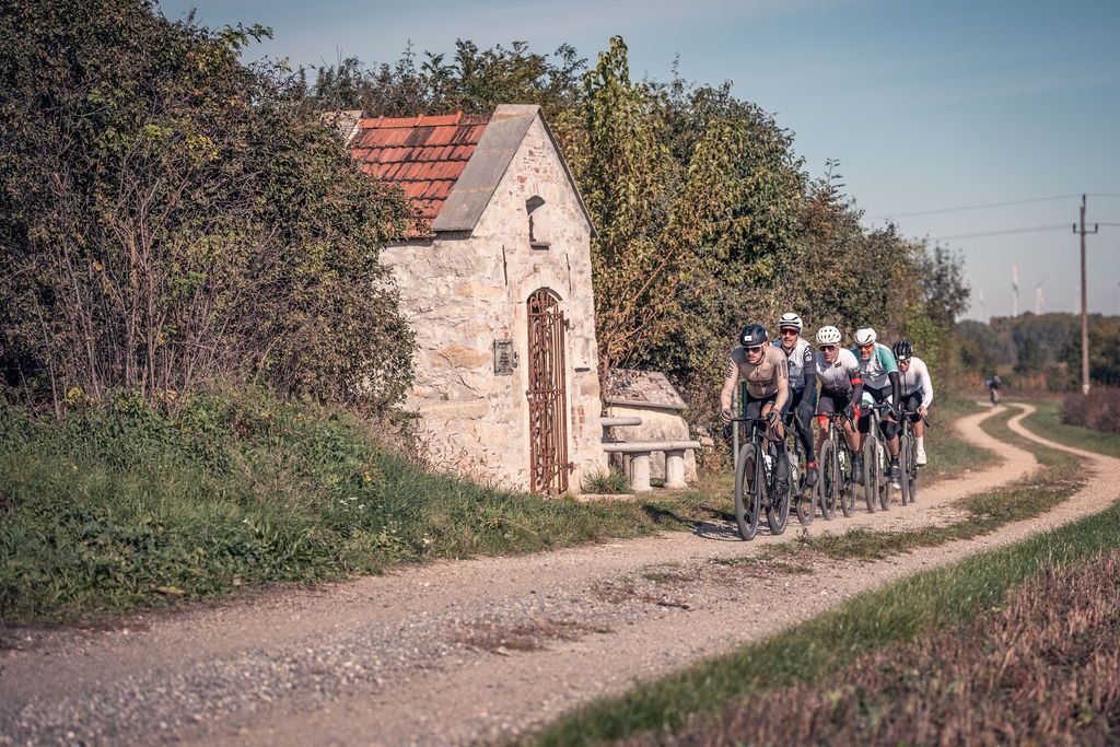 Radfahrer fahren in einer Gruppe auf einem ländlichen Feldweg an einem kleinen Steinhaus vorbei.