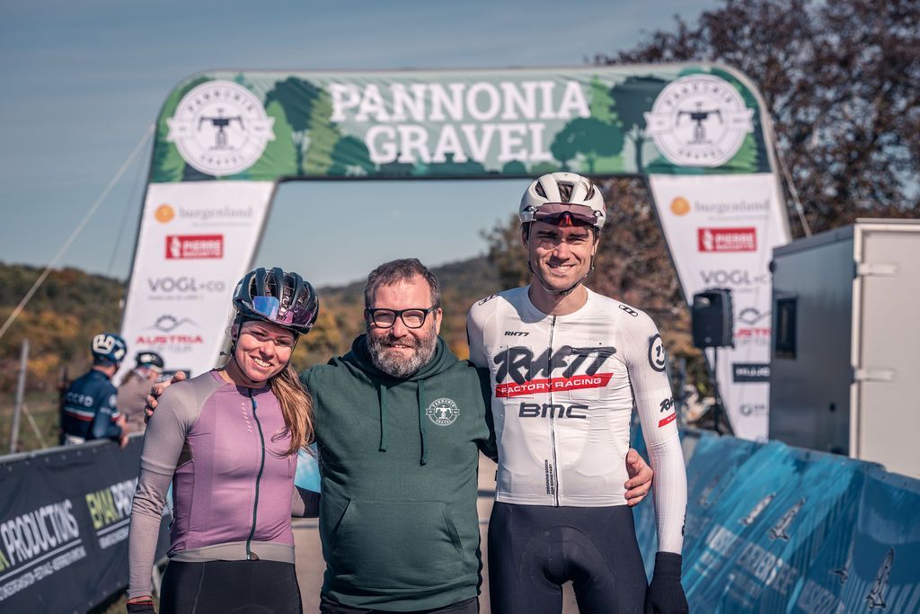 Drei Radfahrer posieren mit Medaillen und einem Lächeln am Zielbogen des Pannonia Gravel-Rennens.