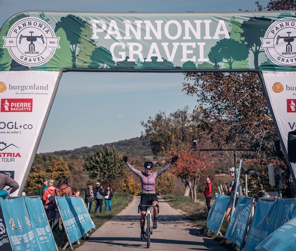Ein Radfahrer überquert die Ziellinie unter einem Bogen aus Pannonia Gravel, die Arme jubelnd erhoben.