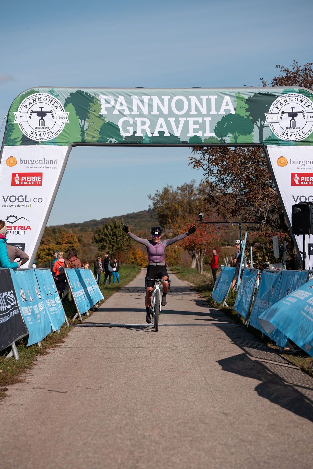 Ein Radfahrer überquert die Ziellinie des Pannonia Gravel-Rennens unter einem grünen Banner, die Arme zum Jubel erhoben.
