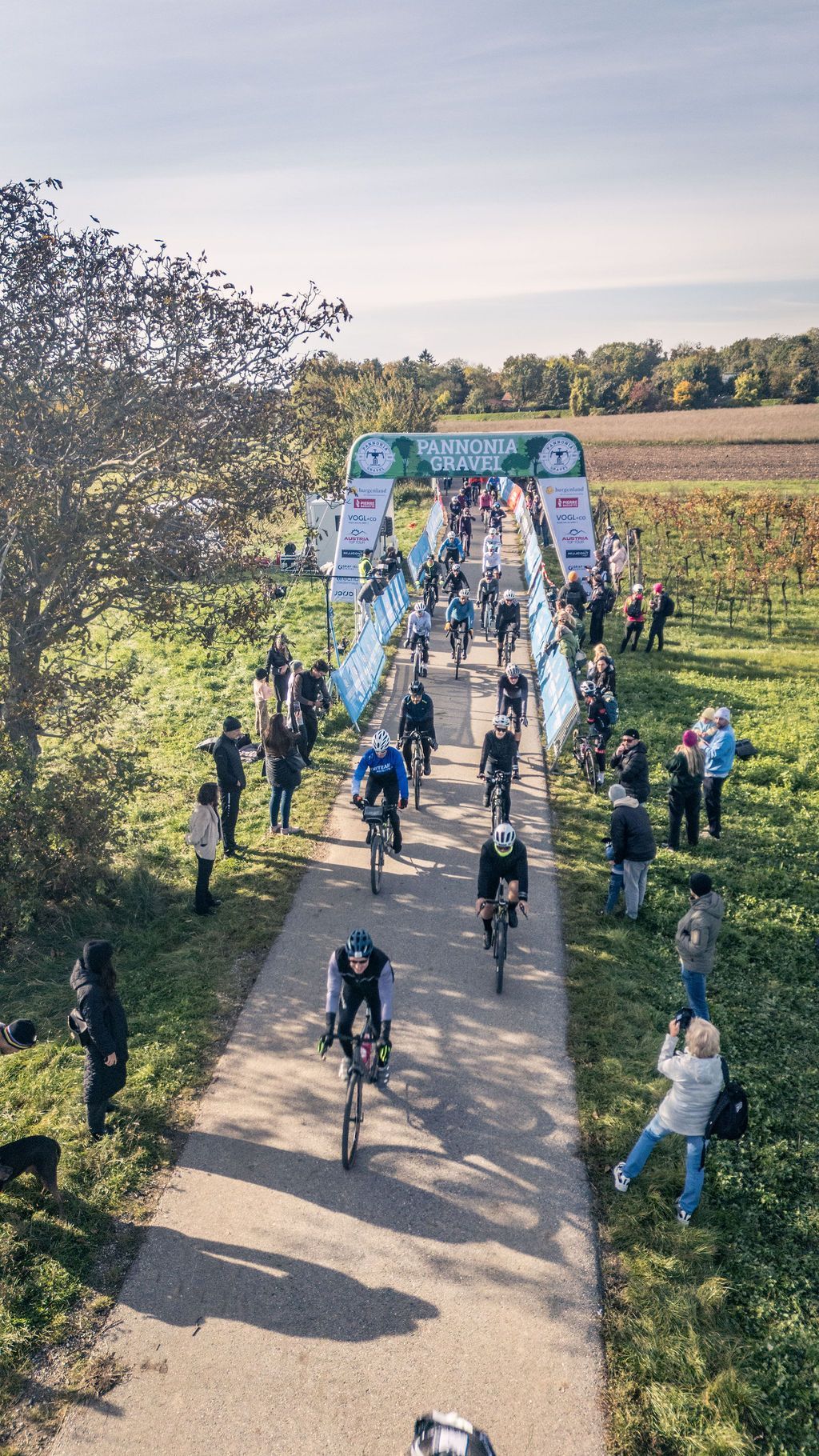 Radfahrer fahren auf einer von Bäumen gesäumten Landstraße auf ein Zielbanner zu, Zuschauer auf beiden Seiten.