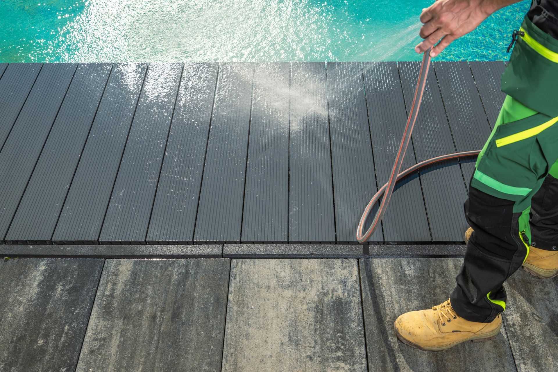 A man is spraying water on a wooden deck next to a pool.