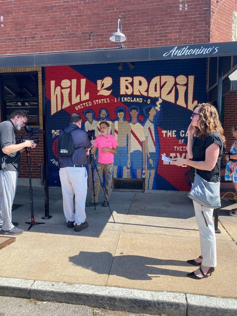 People outside a storefront with a colorful “Hill Brazil” mural, one speaking into a microphone.