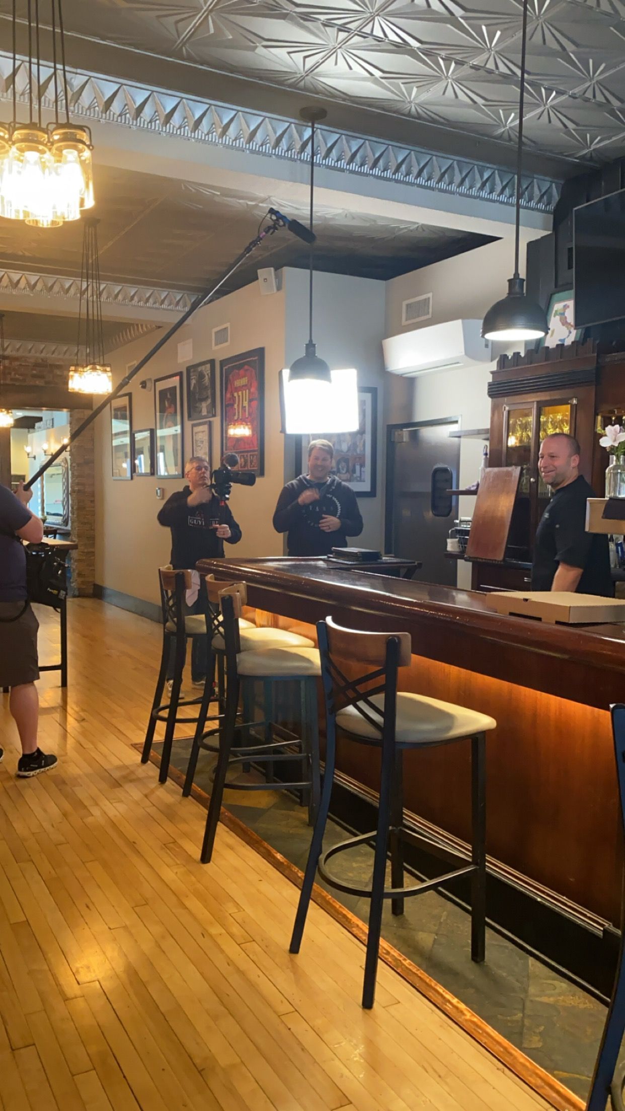 Dimly lit bar interior with wooden counter, stools, hanging lights, and a few people standing near the bar.