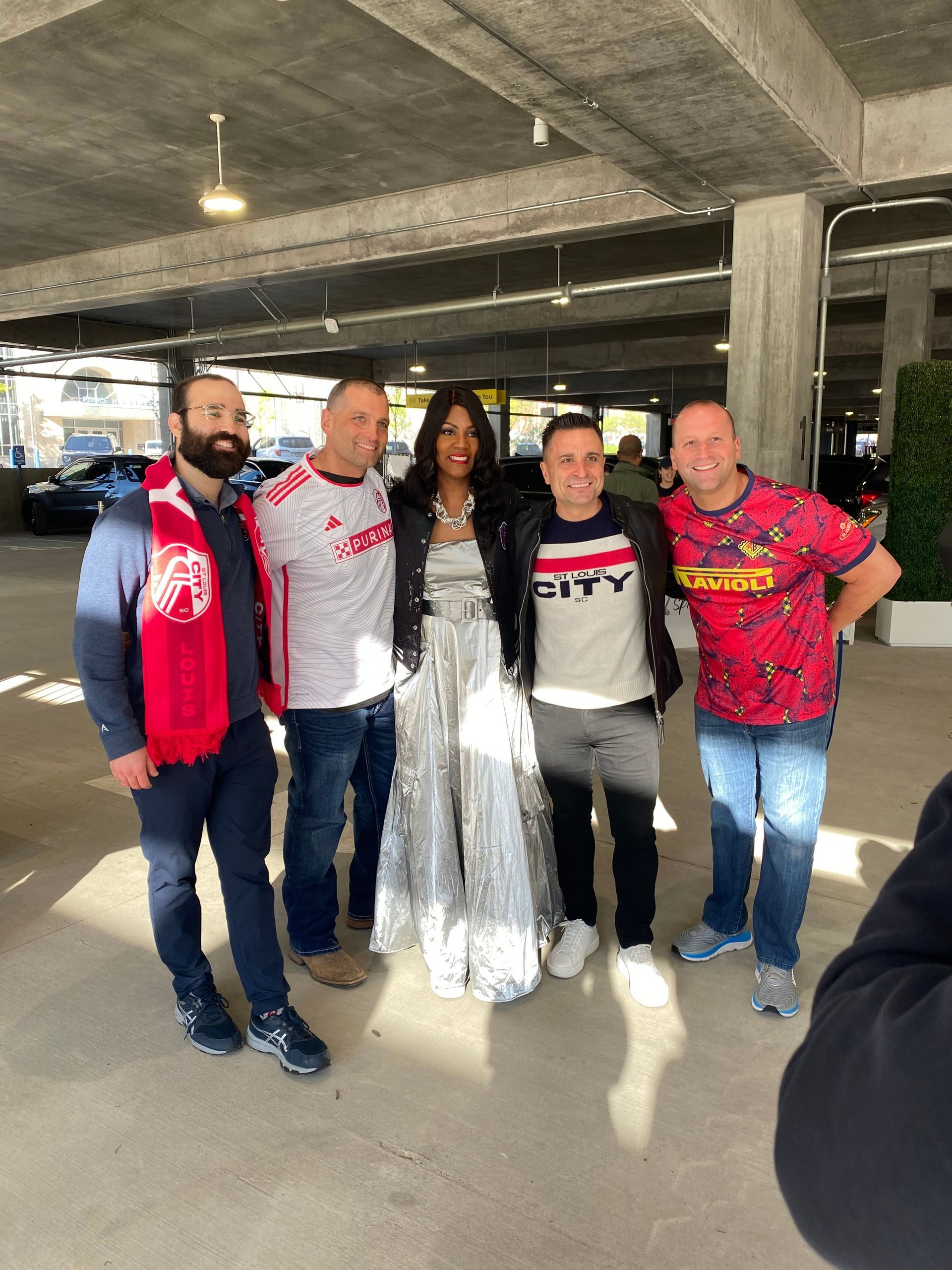 Five people pose together in a parking garage, with one in a white dress and veil-like fabric.