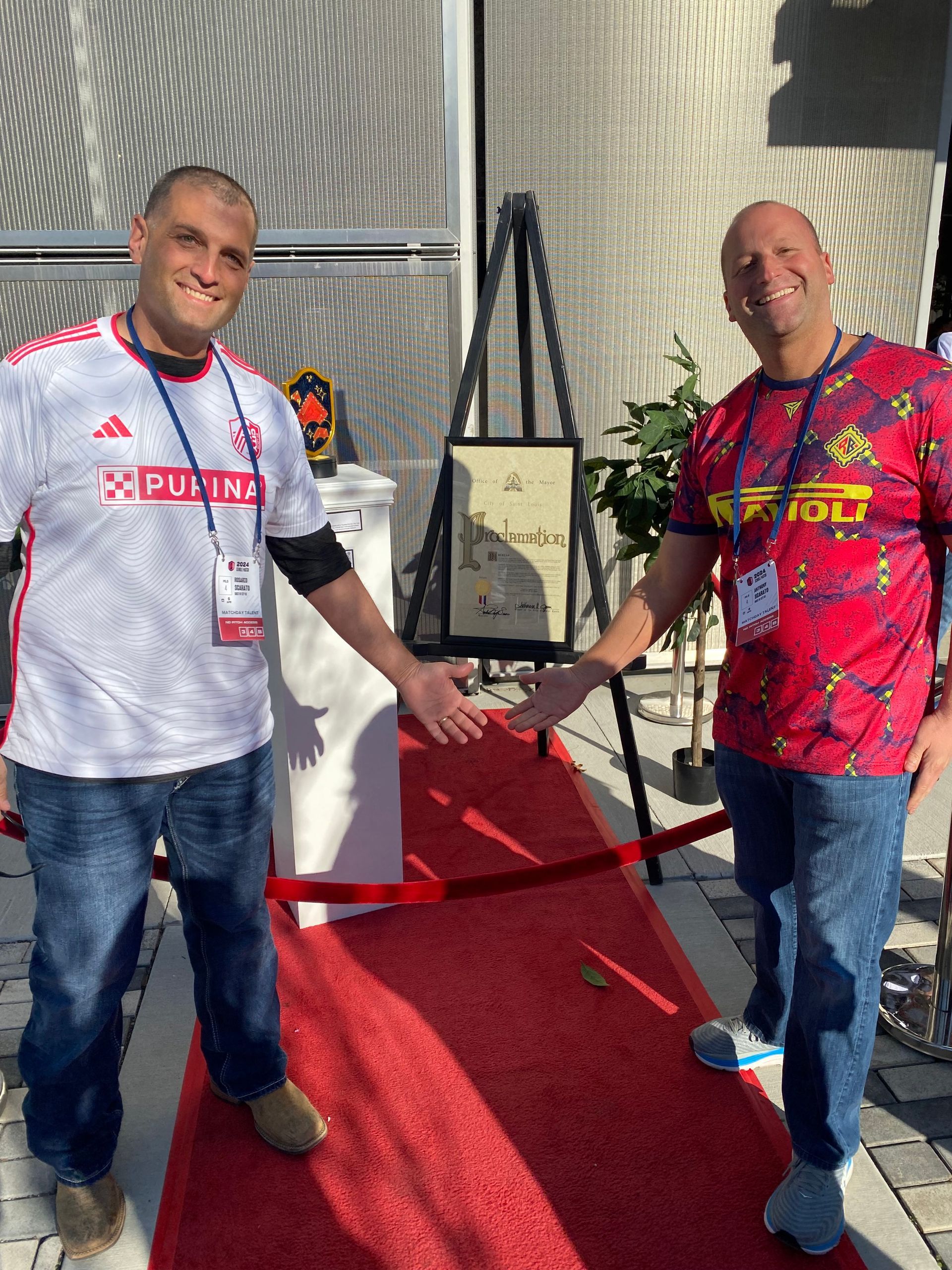 Two men standing on a red carpet, smiling and holding a framed item in a bright indoor setting