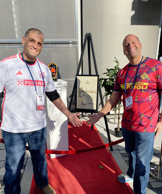 Two men posing on a red carpet beside an easel display, both smiling in colorful event shirts.