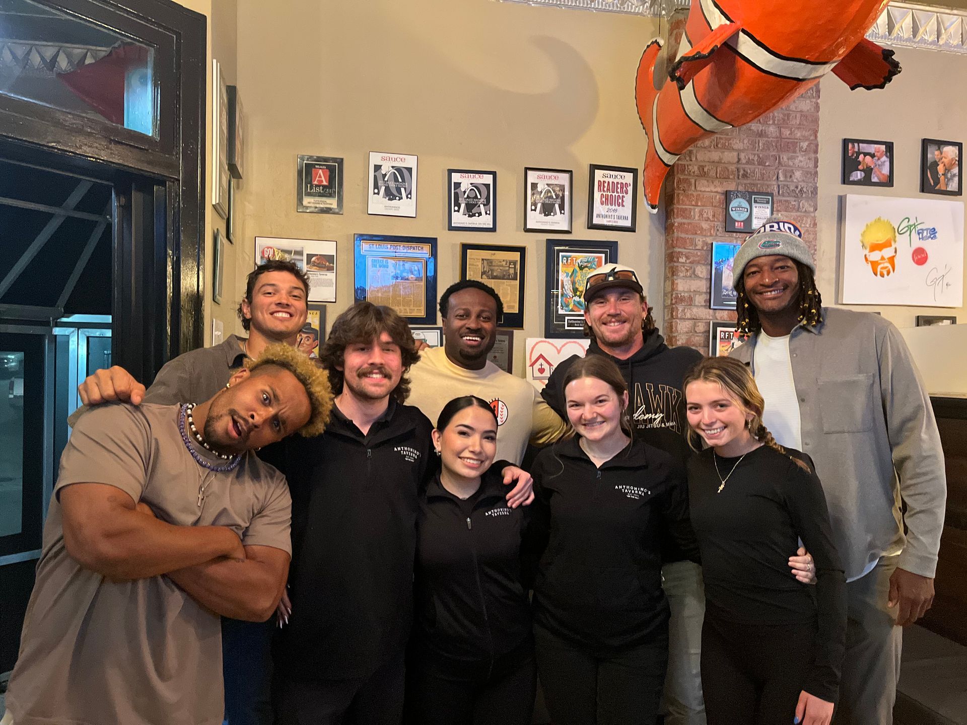 Group of nine smiling people posing indoors at a restaurant, with framed photos on the wall and an orange hanging decoration.