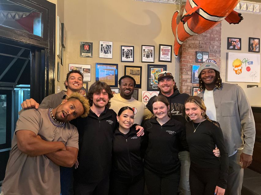 Group of eight people posing and smiling inside a casual restaurant or bar with framed wall art and a large fish decoration.