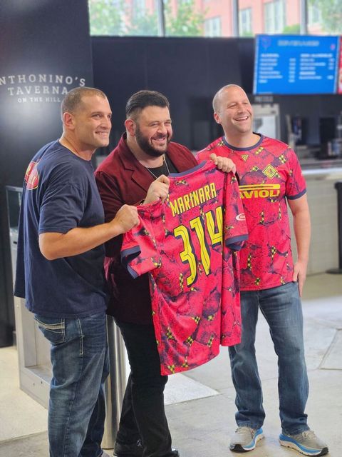 Three men holding a bright pink jersey indoors, smiling for a photo.