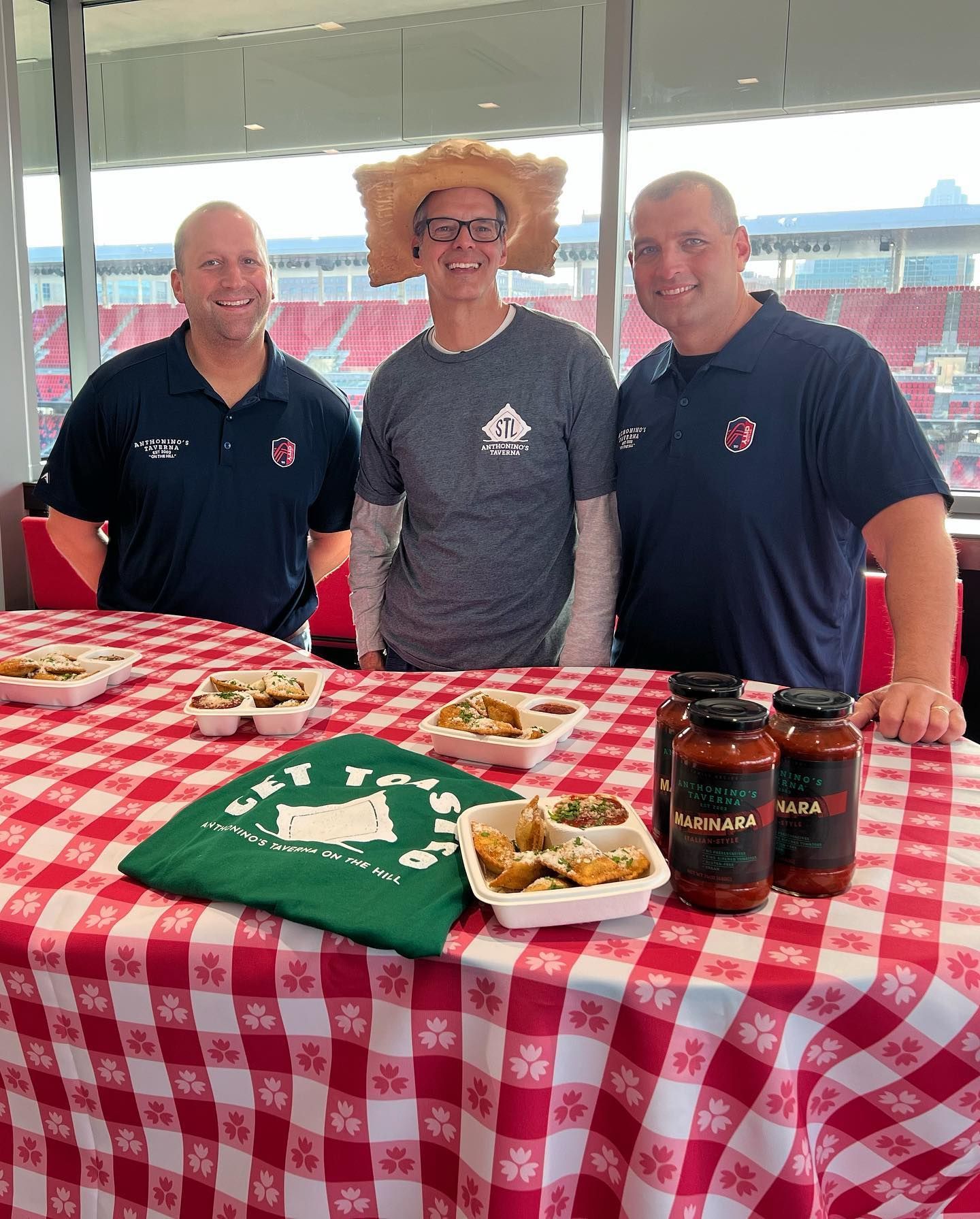 Three people at a sports stadium table with checkered cloth, snacks, green shirt, and drinks.
