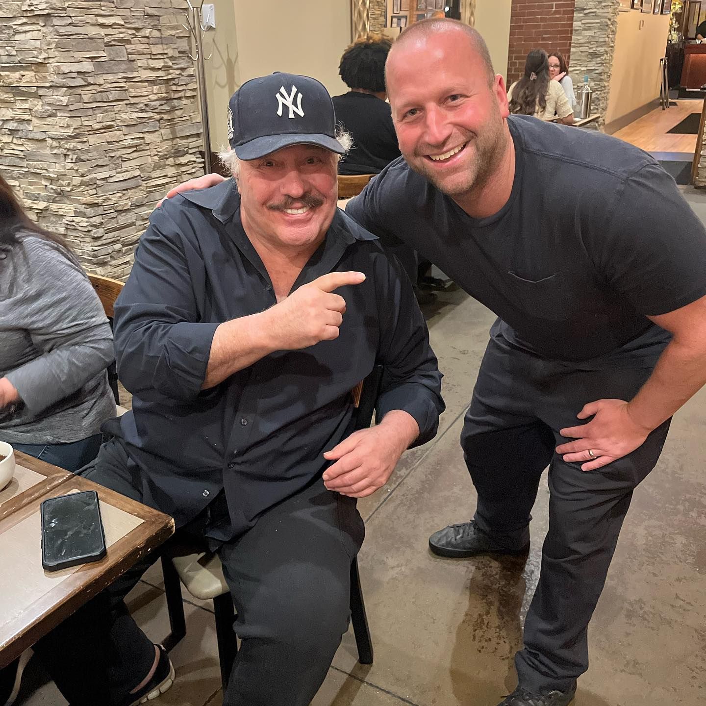 Two men smiling indoors, one sitting and pointing at the other, with stone walls in the background