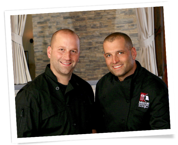 Two men in black shirts smiling indoors in front of a stone wall