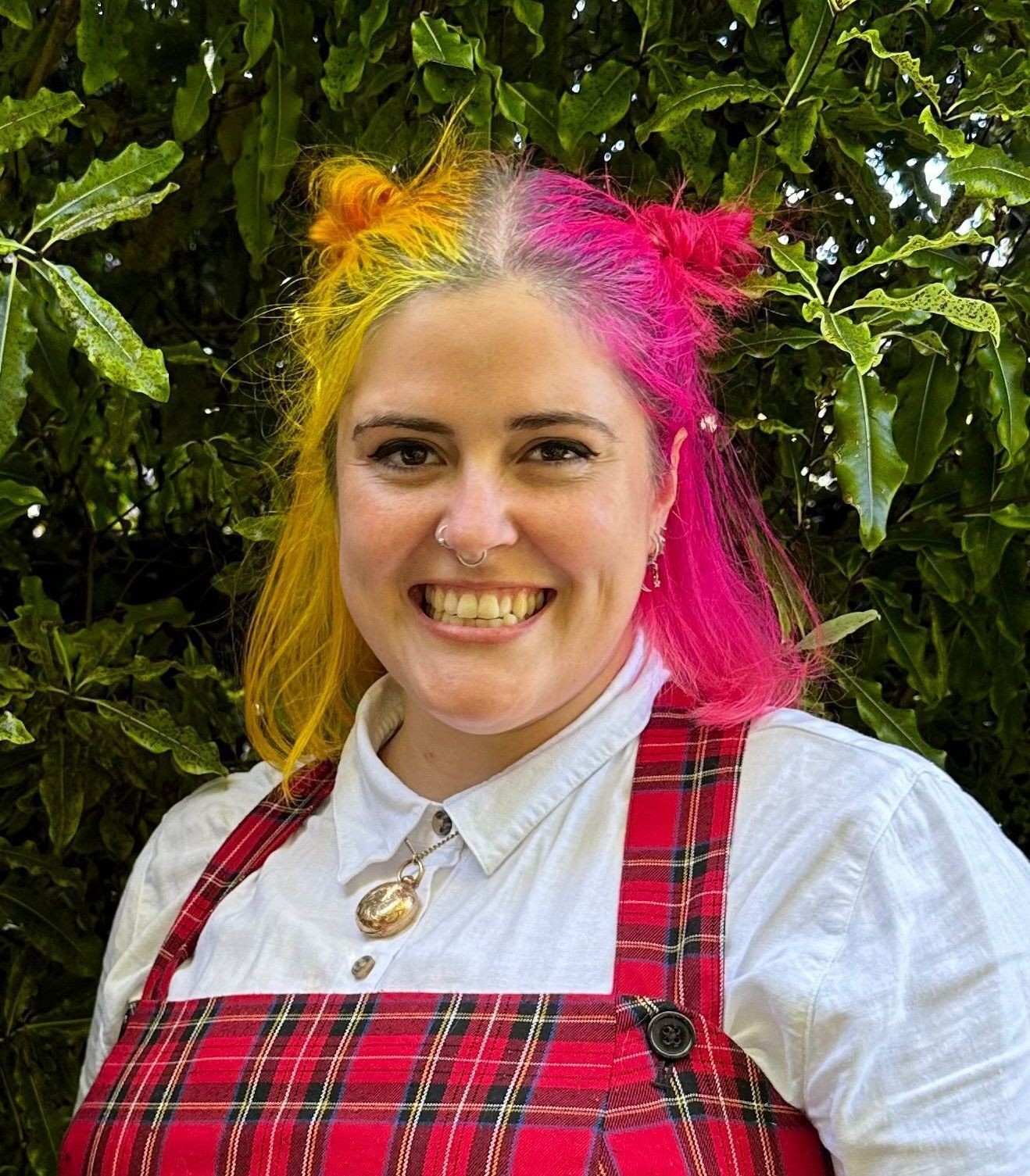 Person with yellow and pink hair smiling, wearing red plaid dress and white shirt, set in front of greenery.