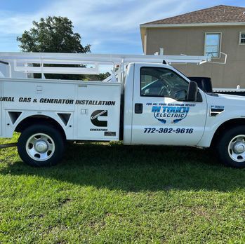 White utility truck with “electric” and gas generator installation branding parked on grass beside a house.