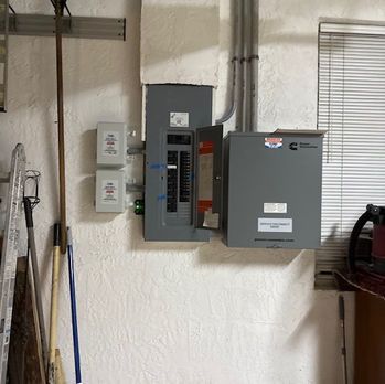 Electrical breaker panels mounted on a white wall in a utility room, with exposed conduit and blinds nearby
