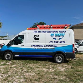 White electrical service van with blue graphics and ladder racks parked on grass under a clear sky.