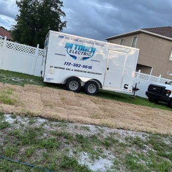 White utility trailer labeled “TJC Electric” parked on a grassy lot beside a fence and house, under cloudy skies