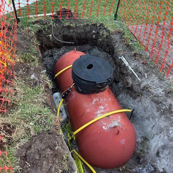 Red underground tank in a dug trench, secured with yellow straps and an orange safety fence nearby.