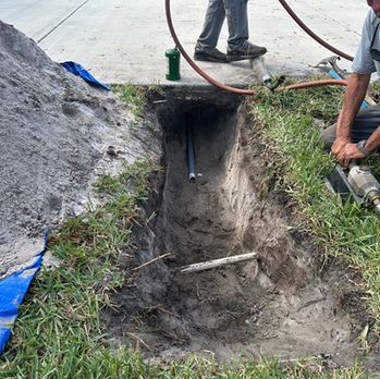Freshly dug trench beside a sidewalk, with workers and hoses around the excavation site.