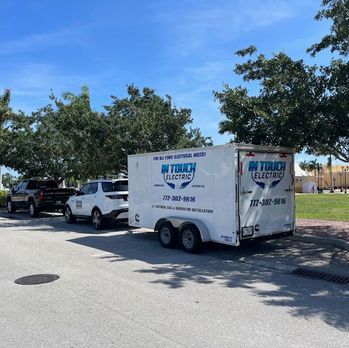 White utility trailer and parked vehicles under trees in a sunny lot.