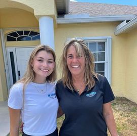 Two smiling women standing outside a yellow house, wearing white and black polo shirts.