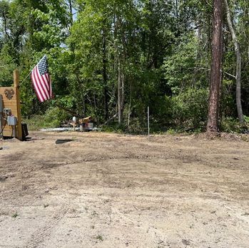 Cleared dirt lot beside wooded area with an American flag hanging near a small shed