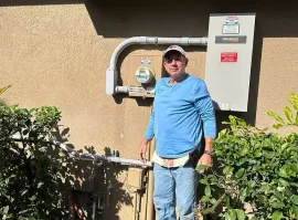 Man standing beside utility meters and a power box on a sunny exterior wall