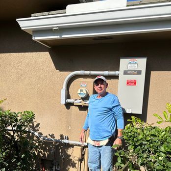 Man standing outdoors beside utility meters and a control panel, wearing a blue shirt and cap.