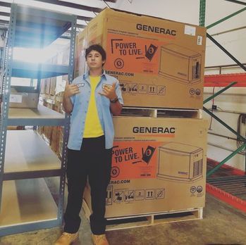 Person standing beside stacked Generac power equipment boxes in a warehouse, giving a thumbs-up.