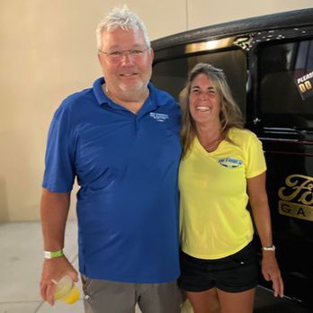 Two people smiling indoors, wearing blue and yellow shirts, standing beside a vehicle.