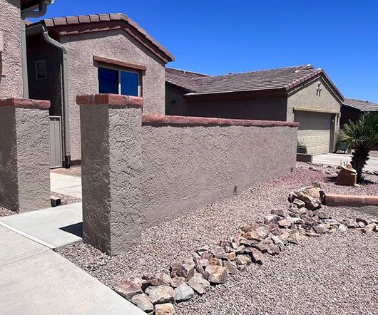 Stucco house exterior with tan walls, red trim, and a gravel yard under a blue sky