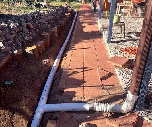 White PVC pipe runs along a brick walkway beside a stone border and patio table.