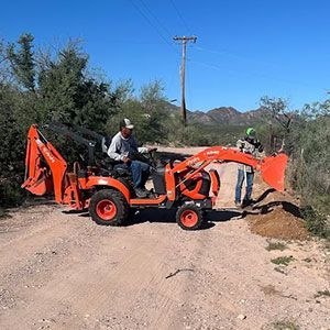 Orange backhoe parked on a dirt road with two workers in a rural area