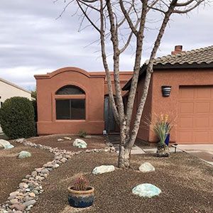 Desert-style house front with terra-cotta exterior, leafless trees, rocks, and a garage under cloudy skies