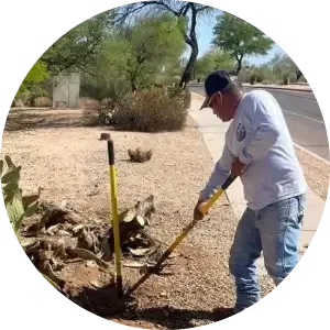 Person digging soil beside a roadside using a shovel, with a yellow post and desert shrubs nearby