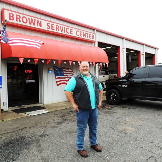 Man standing outside Brown Service Center beside a black truck, with an American flag and red awning.
