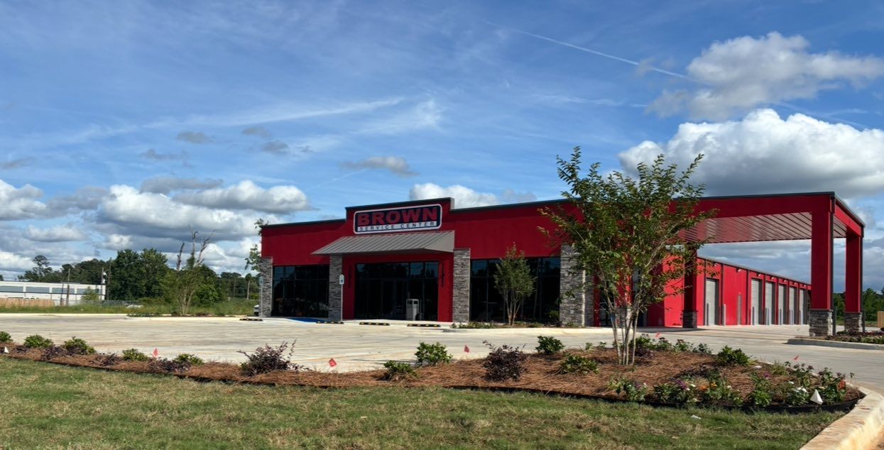 Red building with glass entrance, surrounded by a paved lot, landscaping, and a cloudy blue sky.